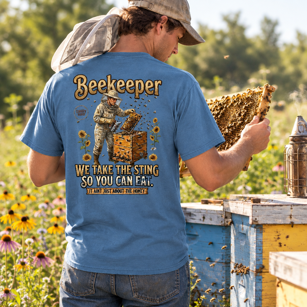 A man wearing a blue graphic t-shirt with a beekeeper design on the back, standing in a sunny field next to beehives and holding a honeycomb frame. The graphic features a beekeeper, sunflowers, and the text 'Beekeeper We Take The Sting So You Can Fail.