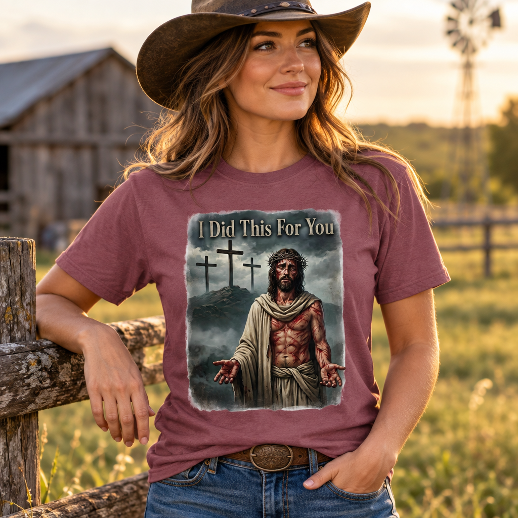 A woman wearing a brown cowboy hat and a maroon graphic t-shirt with the text 'I Did This For You' and an illustration of Jesus Christ, posing outdoors in a rural setting with a wooden fence and barn in the background.